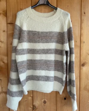 Wit-beige gestreepte trui van Emi-Jo met subtiele glitterdetails. Zacht, cosy en trendy – de perfecte knit voor herfst en winter.