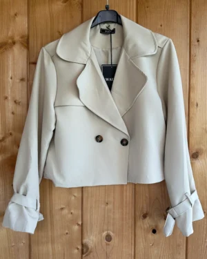 Stijlvolle beige cropped trenchcoat van Wauw Fashion. Met dubbele knopen en ceintuurdetails bij de mouwen. Tijdloos, elegant en veelzijdig te combineren.